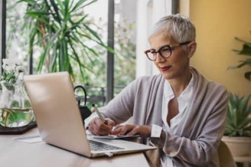 A woman sits at a table looking at a laptop as she writes in a notebook.