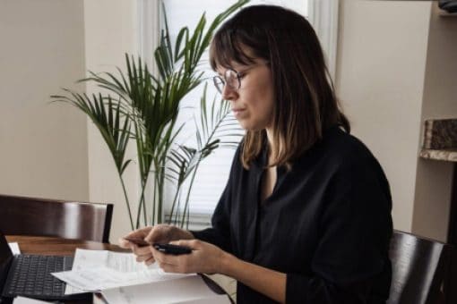 A woman sits at her kitchen table with her laptop and reviews her paper credit card statements.
