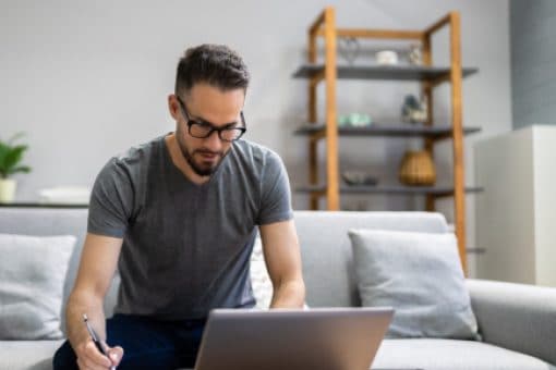 A man with glasses sits in front of his computer while taking notes.