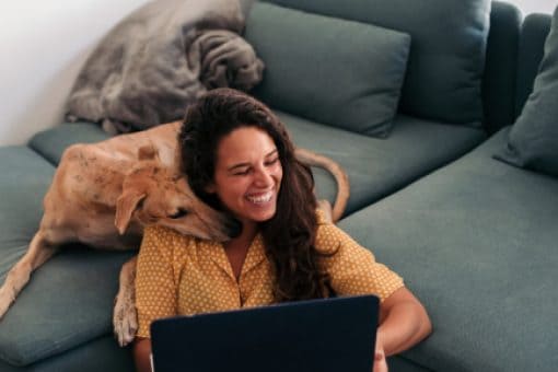 A young woman sitting on the floor and holding a laptop laughs as a dog nuzzles her neck.