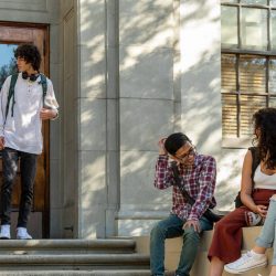 College students are standing and sitting outside of a building.