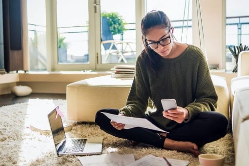 Young woman sits cross-legged on floor holding mobile phone and papers.