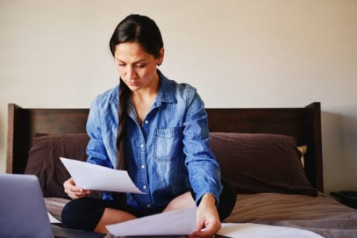 A woman in a blue shirt sits on her bed looking at a paper bill.