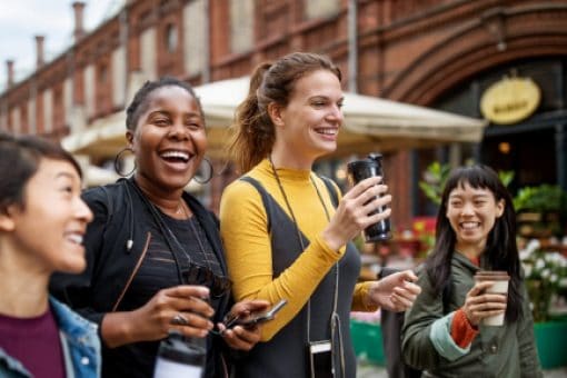 Young women walk down the street laughing and drinking coffee.