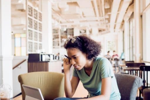 Young woman sits on a coffee shop chair working on a laptop.