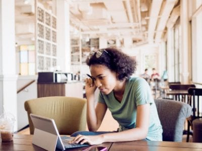 Young woman sits on a coffee shop chair working on a laptop.