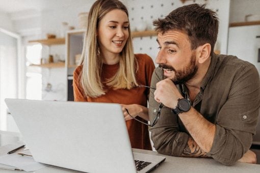 Young couple smiles together as they look at a computer screen in their kitchen.