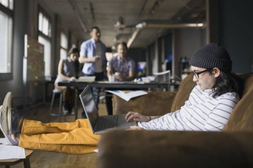 A man works on his laptop in an open office with coworkers grouped in the background.
