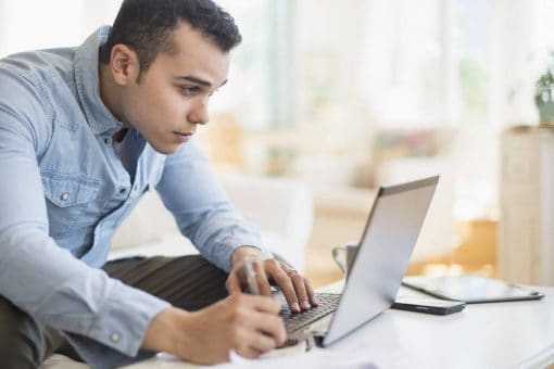 A man sitting on a couch reviews his credit card statement on his laptop.