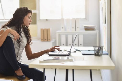 Young woman sits casually at her desk looking at her laptop.