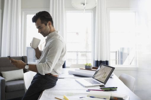 A young man leans against his desk and looks at his phone.