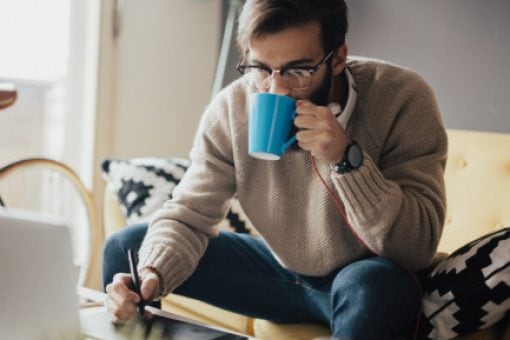 Young man sips coffee while using digital stylus on tablet.