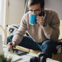 Young man sips coffee while using digital stylus on tablet.