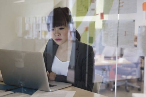 Woman stares at her laptop screen behind glass walls in an office.