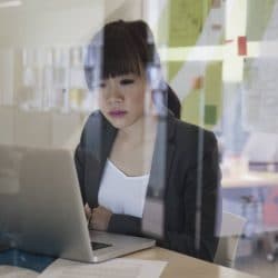 Woman stares at her laptop screen behind glass walls in an office.