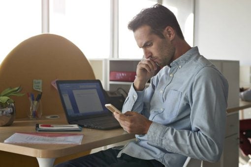 Man sits at a desk in his office, looking down at his cell phone.
