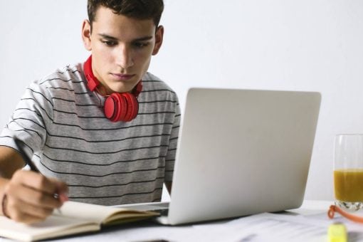 Young man in a black striped shirt wears red headphones around his neck while writing in a notebook.