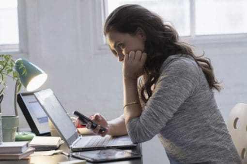 Woman sits at a desk looking intently at her laptop while holding her mobile phone.
