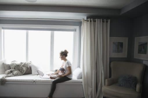 Mom with infant sits on windowsill looking at a laptop.