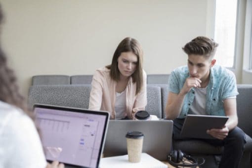 College students sit together in a common room while working on laptops and a tablet.