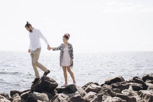 A couple walks on rocks near the ocean while traveling.