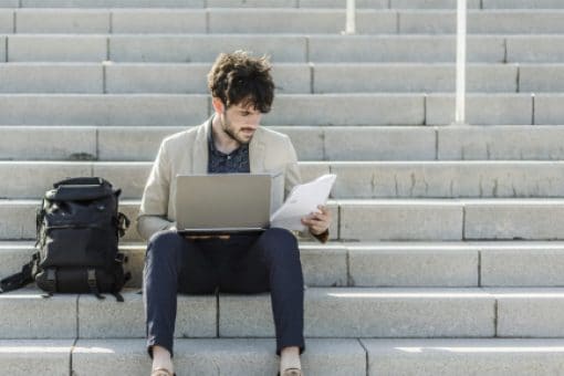Young man on steps looking at laptop and papers