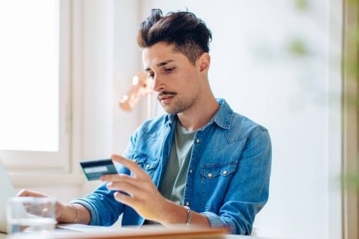 Young man holds his credit card in one hand as he sits at a desk with a laptop