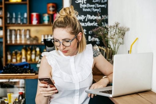 A woman sits at laptop in a café and checks her phone.