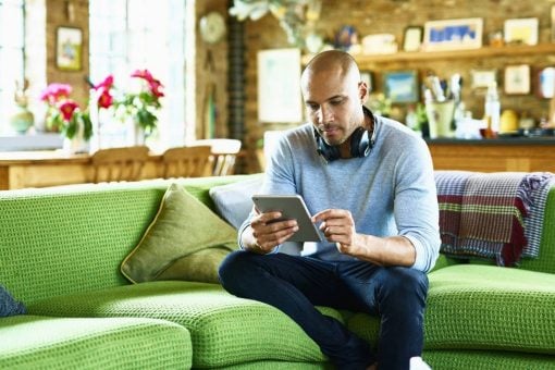 Man with headphones hung around his neck sits on a couch looking at a digital tablet.