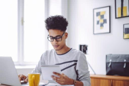 Young man reviews notebook as he works on laptop.