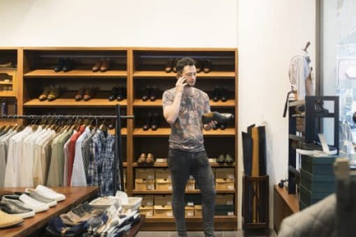 Man in a clothing store holds a shoe to try on while on his mobile phone.