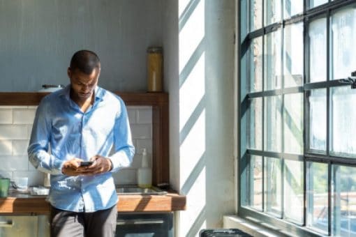 A man leans against his kitchen sink looking down at his phone.