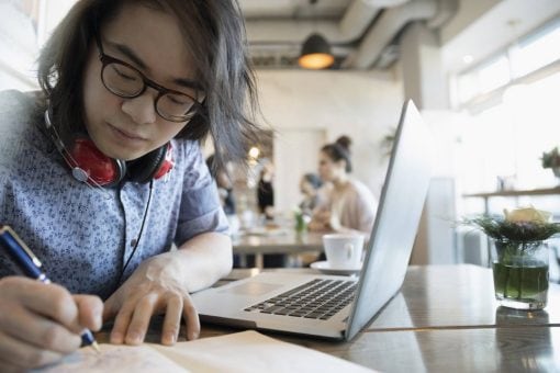 A young man takes notes in a notebook next to his laptop.