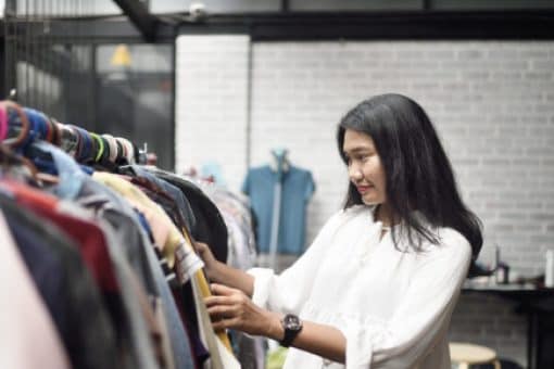 A woman with dark hair wearing a white top looks through clothing rack in a boutique.