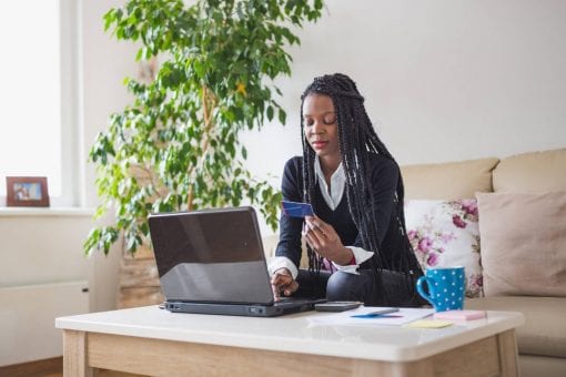 Young woman sits on her couch working on a laptop and holding a credit card in her hand.