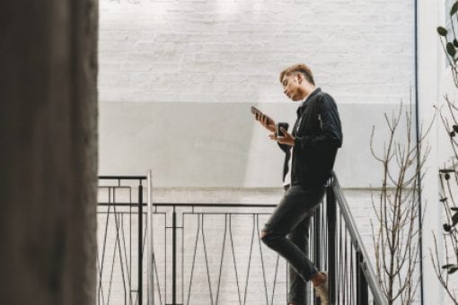 A young man stands in stairwell holding paper coffee cup and scans his mobile phone.