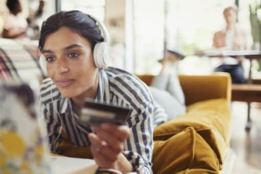 Young woman lays on her couch holding credit card and looking at laptop.