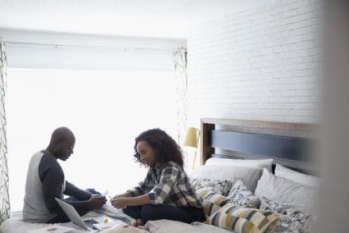 A man and a woman sit on a bed with a laptop and papers spread out beside them.