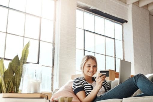 Young woman reclines on couch holding a secured credit card and digital tablet.