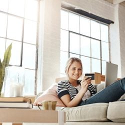 Young woman reclines on couch holding a secured credit card and digital tablet.