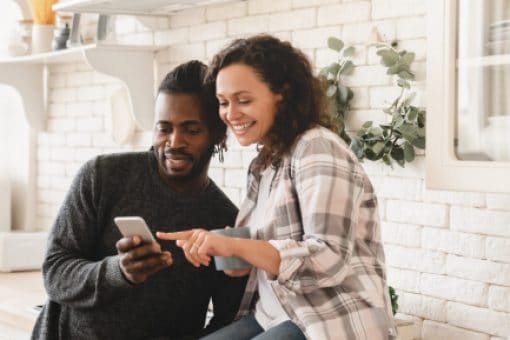 A couple smiles in their kitchen as they look at the man’s phone