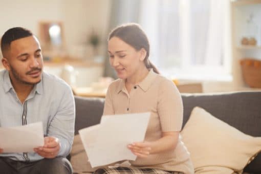 A man and woman sit on couch looking at paper documents.