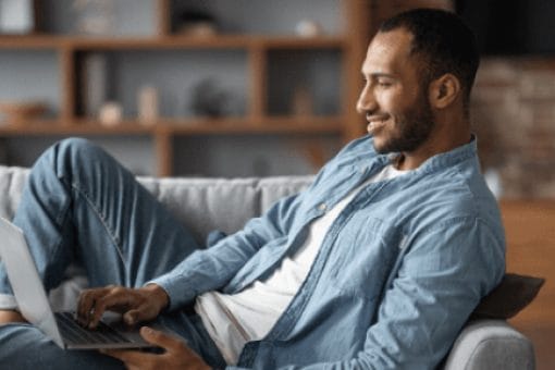 Man in blue shirt smiles looking at laptop as he reclines on a couch.