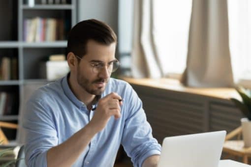 A man wearing a blue dress shirt looks at his laptop with a serious expression.