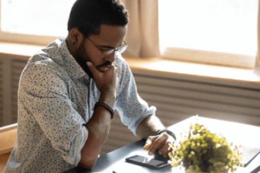 Man sitting at a desk in a sunny room looks down thoughtfully at his phone. A closed notebook sits on the desk.