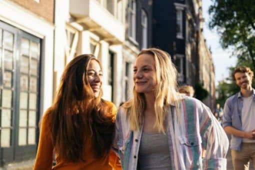 Two young women laugh and talk while walking down a busy sidewalk in a shopping district.