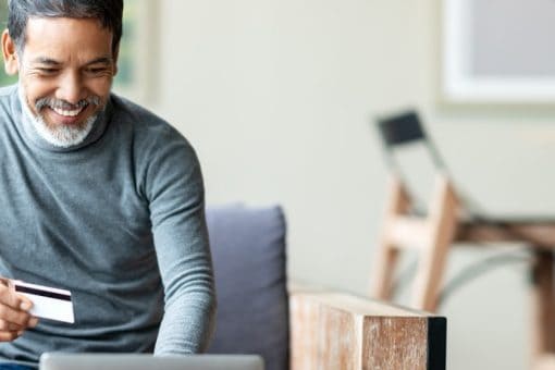 An older man refers to hisholds a credit card to manage his credit card account online on hisand smiles as he looks at a laptop.