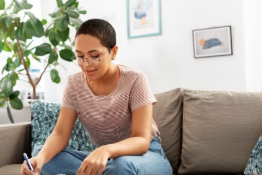 A young woman sits on her sofa and works on calculating her income with pen and paper.