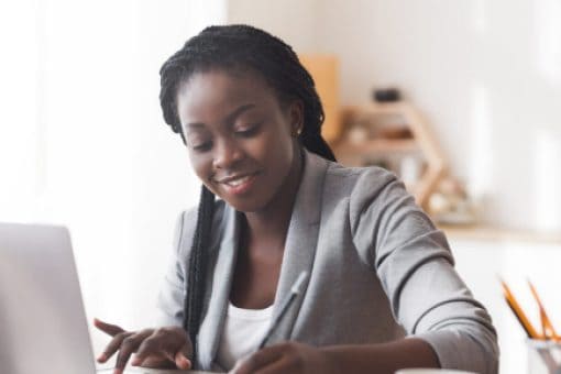 Woman sitting at a desk on a laptop writing in a notebook and smiling.