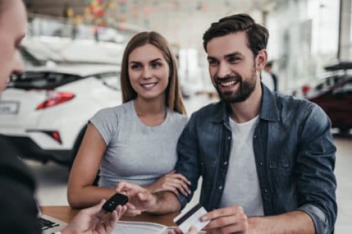 A young couple sits at a table in a car showroom, hands their credit card to a dealer while receiving a new car key.